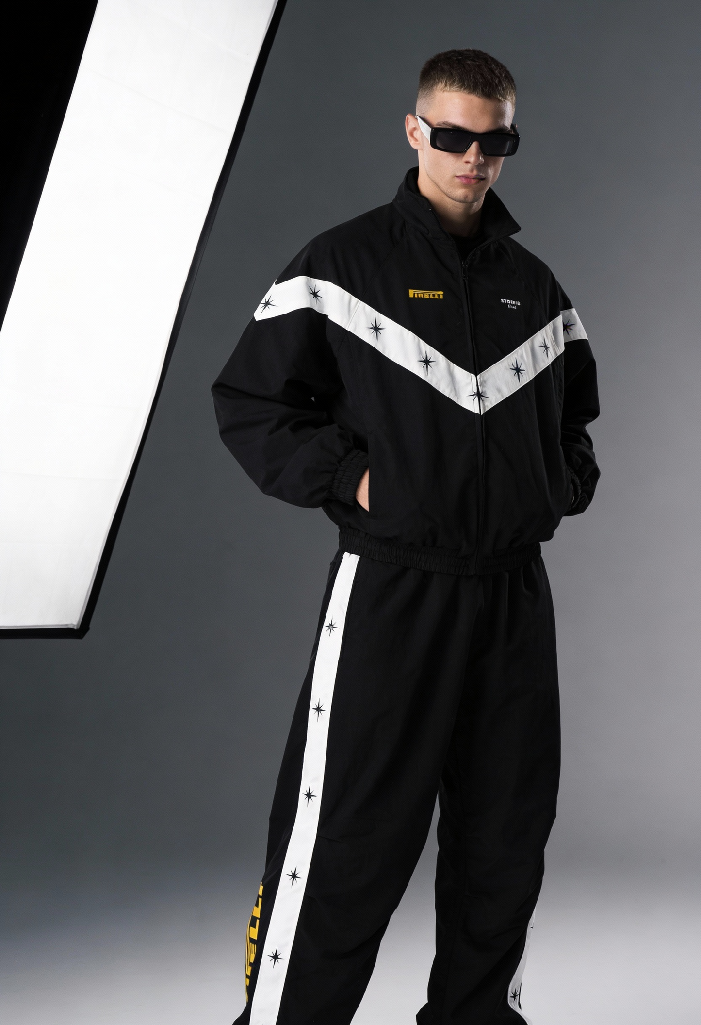 Person wearing a black tracksuit with white stripes and sunglasses on a gray background