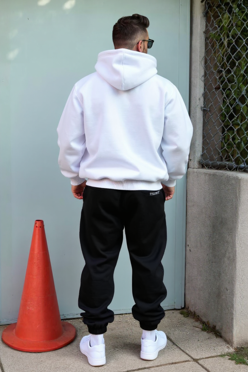 Person wearing a white hoodie and black pants standing next to an orange traffic cone against a light blue wall.